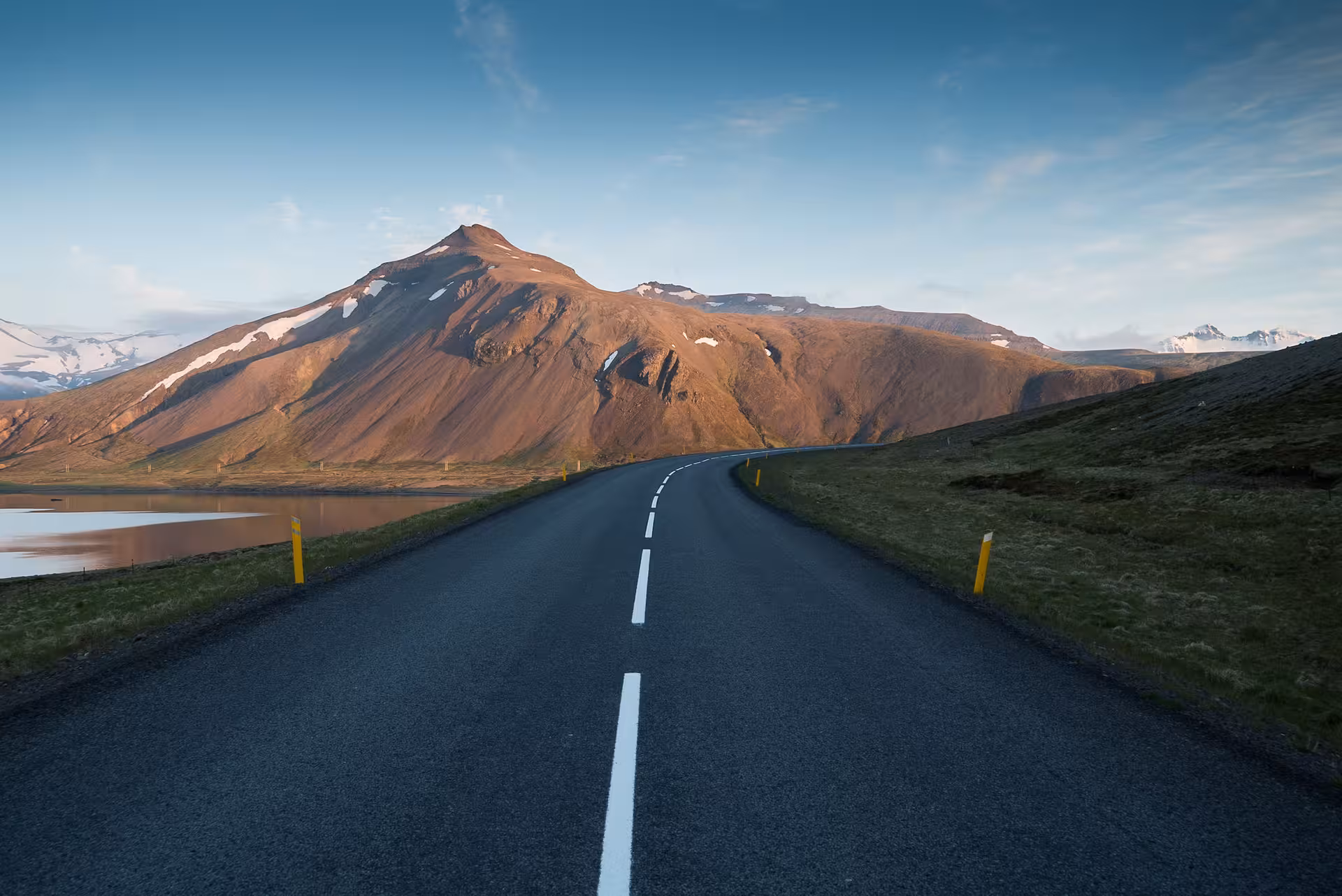 Narrow road leading toward Snæfellsnes volcanic mountain with snow patches and a calm lake on the left