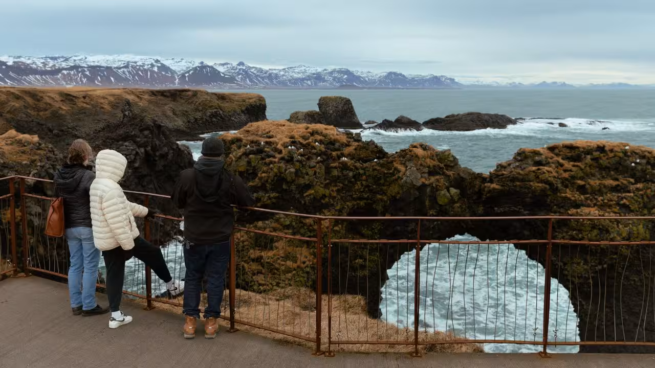 Three tourists at railing overlooking rocky Snaefellsnes coastline with snow-capped mountains beyond