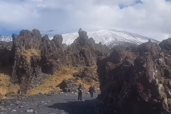 Hikers walking among volcanic rock formations with snow-capped Snæfellsjökull volcano in background