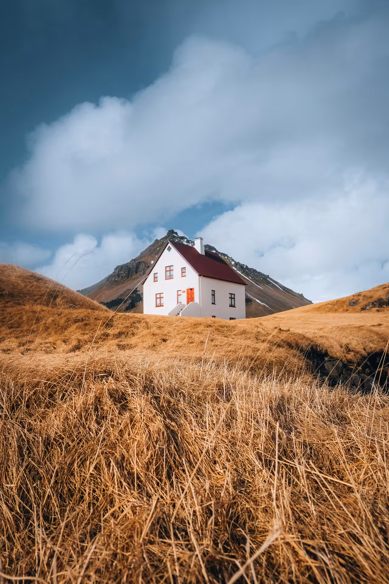 White house with red roof nestled among golden grass hills below a dramatic mountain peak in Snaefellsnes Iceland