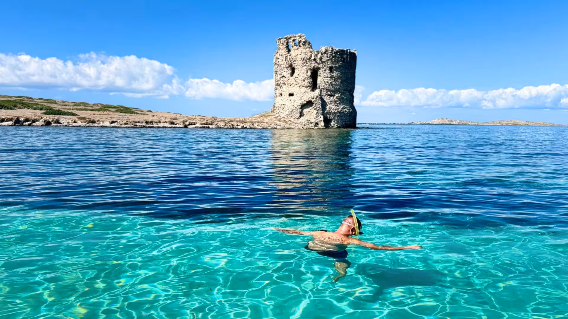 Snorkeler enjoying vibrant waters near Asinara, with historic coastal tower and clear blue skies in the background.