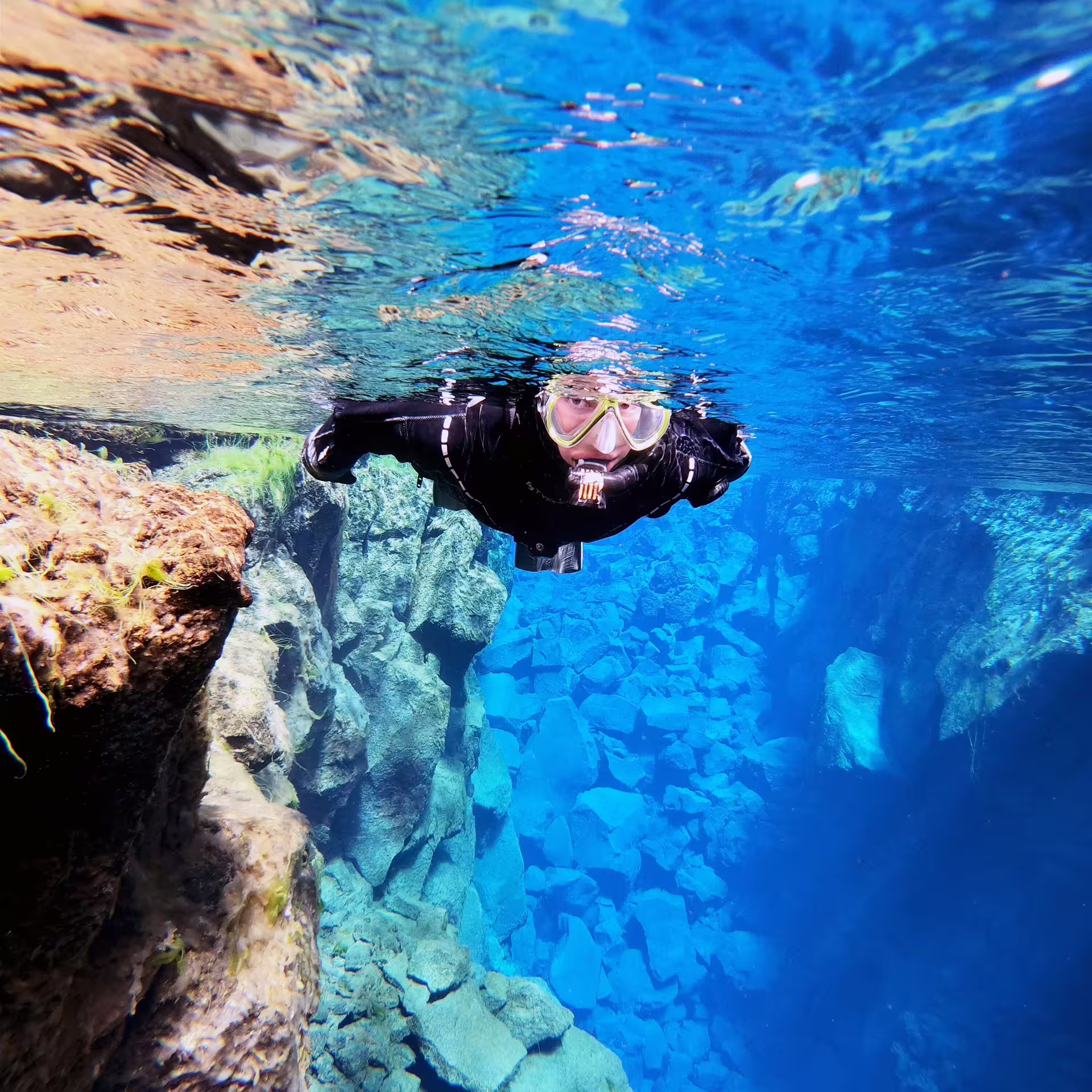 Snorkeler in wetsuit explores the crystal-clear waters of Silfra, Iceland, with stunning underwater rock formations visible.