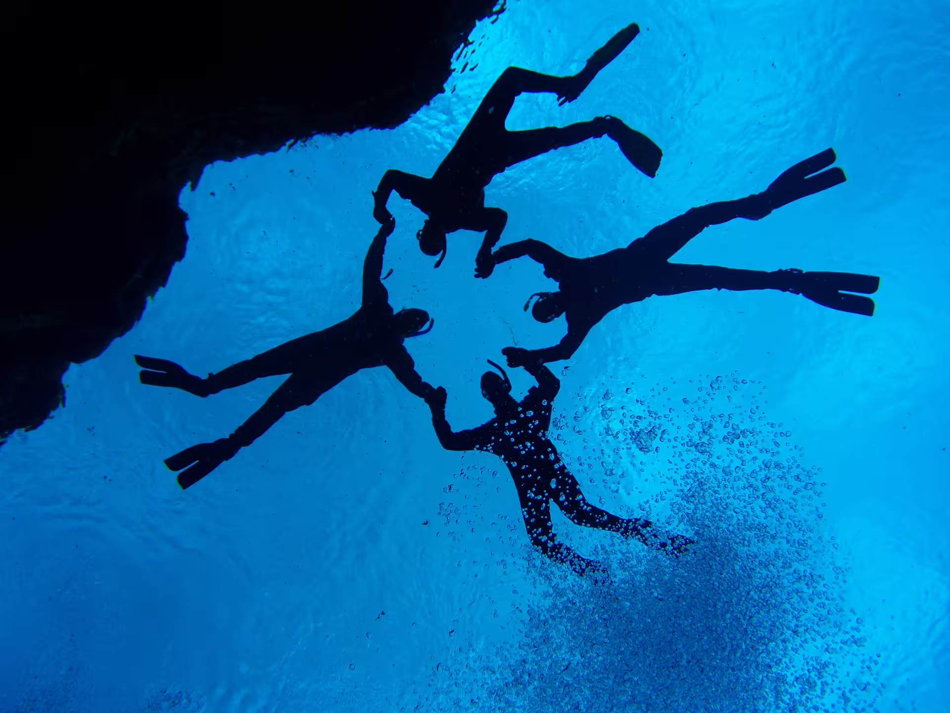 Silhouettes of four scuba divers swimming underwater against vivid blue water with air bubbles rising