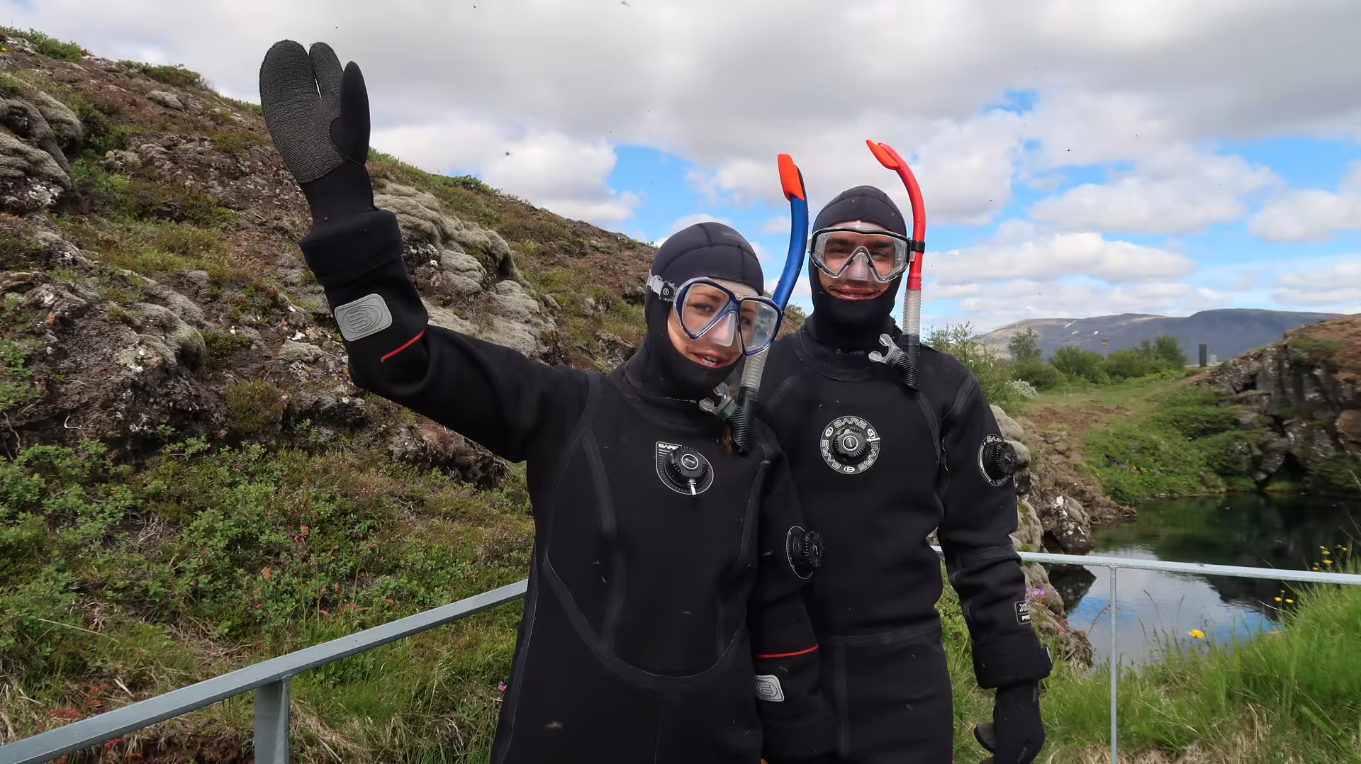 Two snorkelers in black drysuits and masks waving outdoors in Iceland's rocky landscape