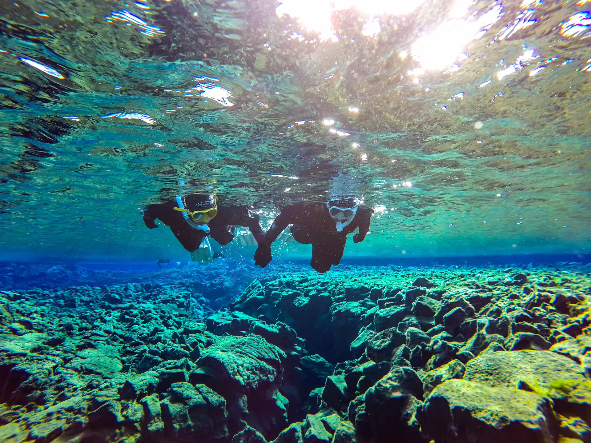 Two snorkelers in drysuits floating above volcanic rock fissure in crystal clear blue water at Silfra, Iceland
