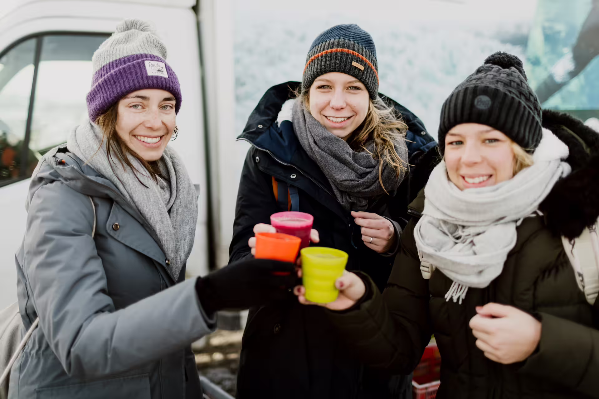 Three smiling women in winter gear toasting with colorful cups outdoors in Iceland