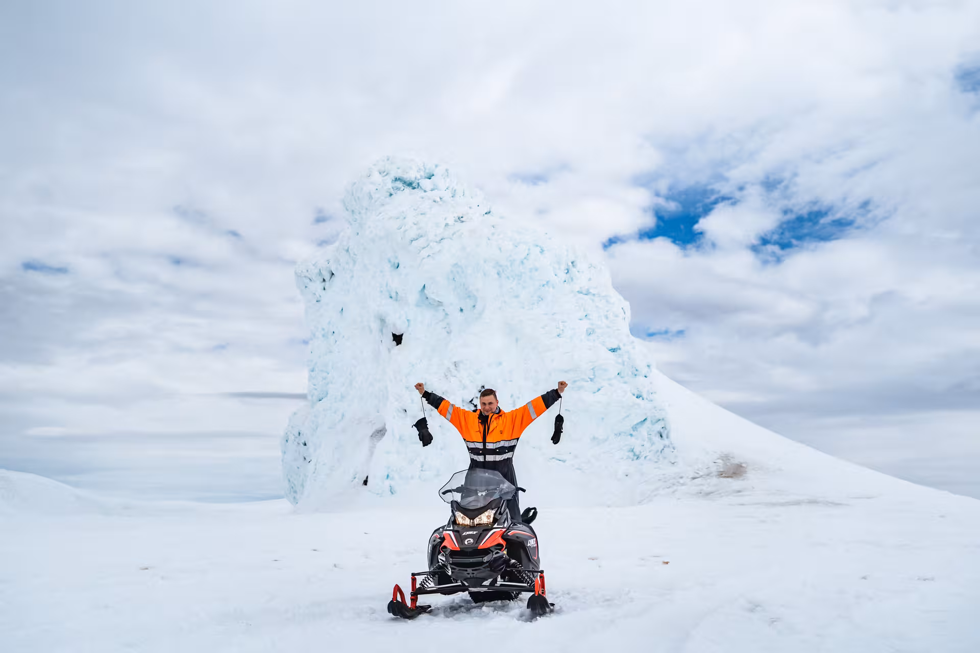 Guide in orange jacket celebrating on snowmobile in front of glacial ice formation on Eyjafjallajokull