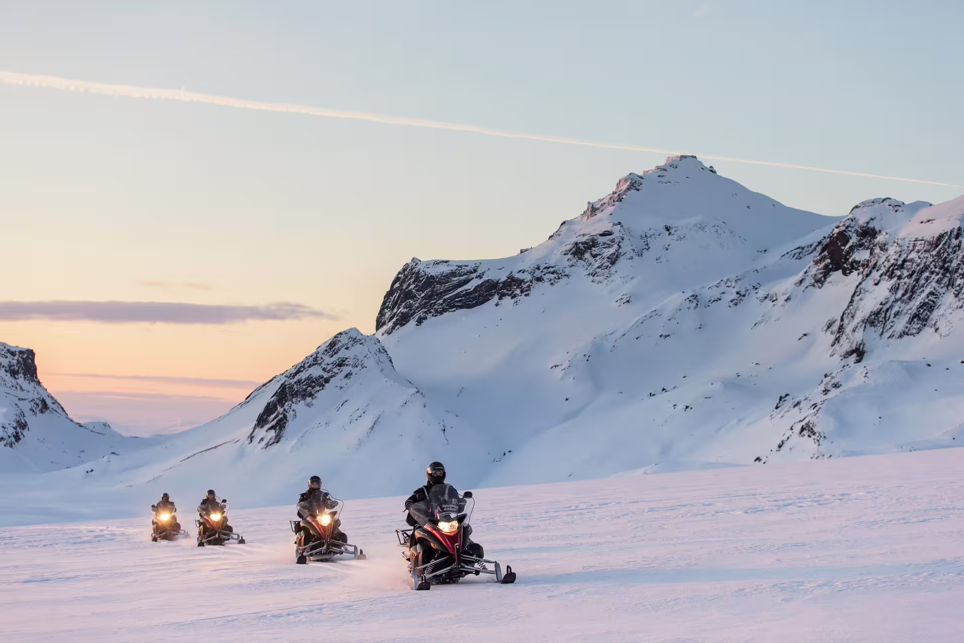Group of snowmobilers riding across a vast snowy glacier with dramatic snow-capped mountains at sunset in Iceland