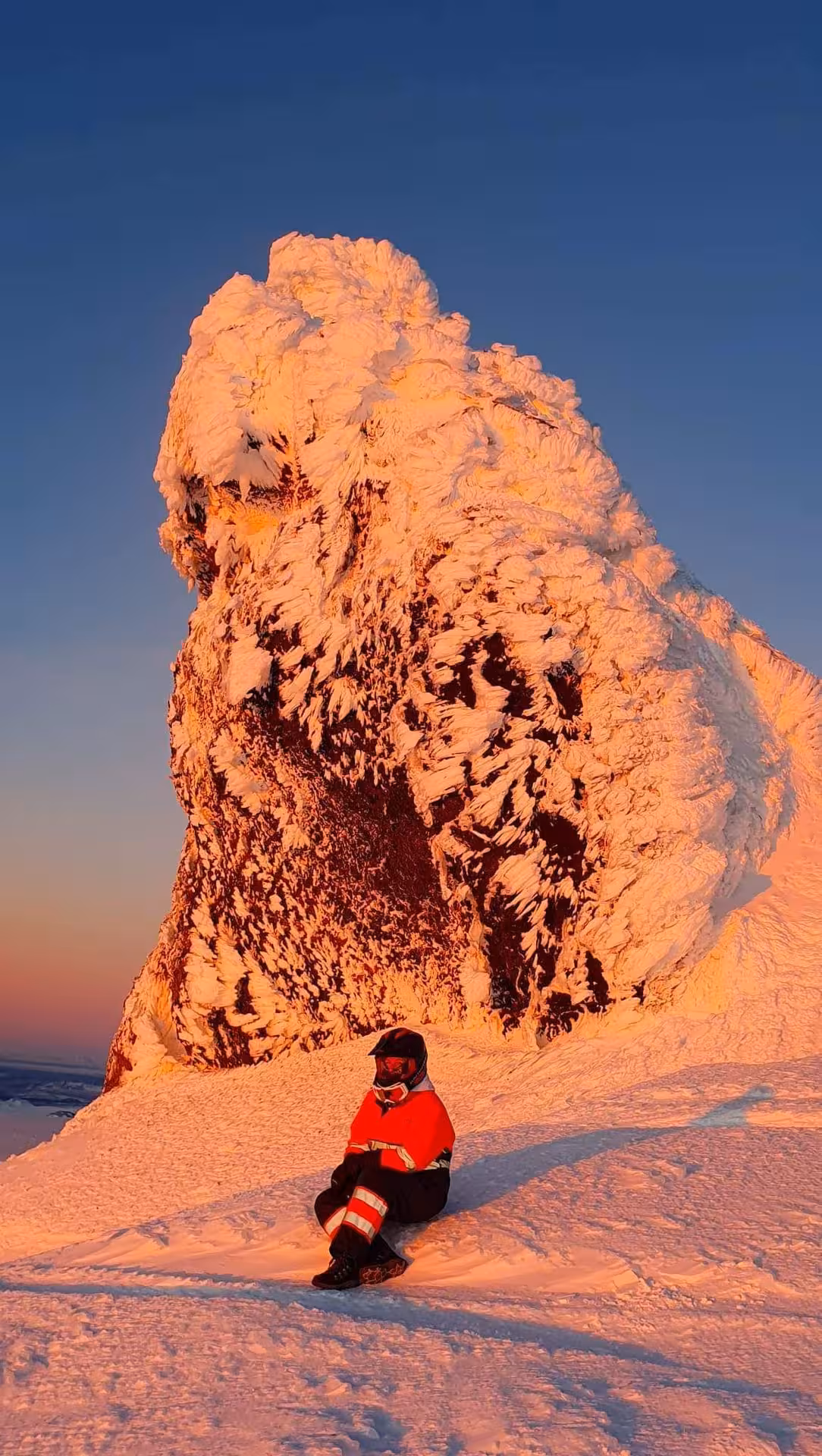 Snowmobiler in red gear resting beside massive snow-covered rock formation on Eyjafjallajokull at golden sunset