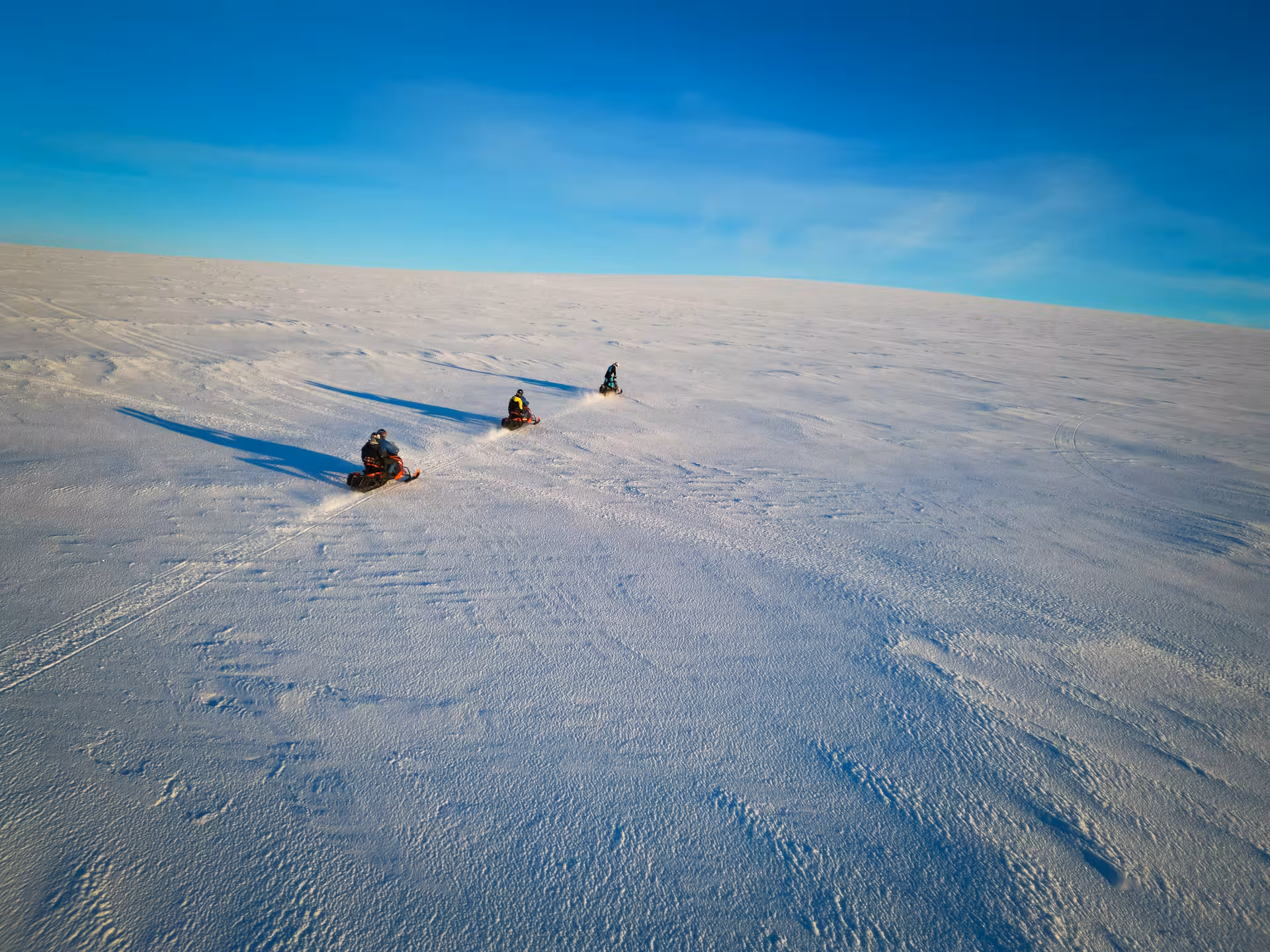 Three snowmobilers riding across Langjokull glacier under a clear blue sky in Iceland