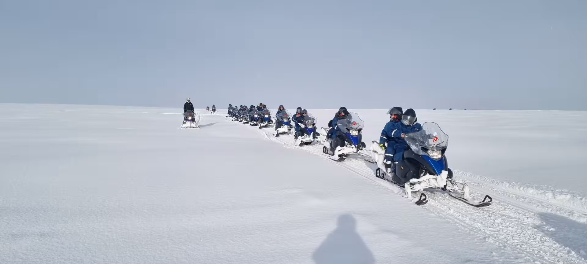 A group of snowmobilers riding in a line across a vast snowy glacier on a Golden Circle super jeep and snowmobile tour from Reykjavik Iceland.