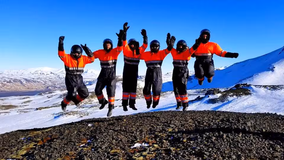 Six snowmobilers in orange safety suits jumping joyfully on snowy Landmannalaugar highlands terrain
