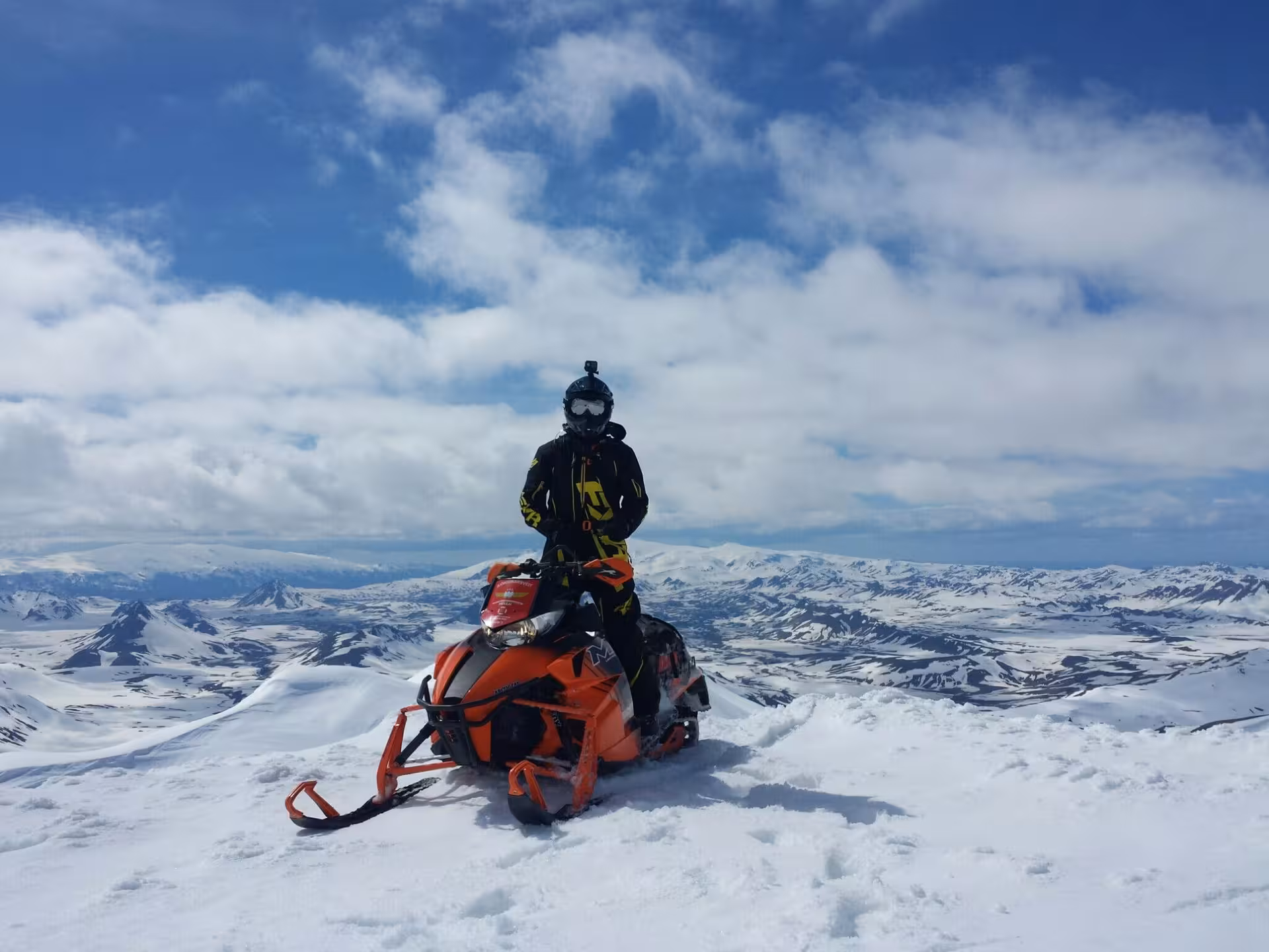 Rider on orange snowmobile atop snowy Landmannalaugar Highlands with panoramic mountain views