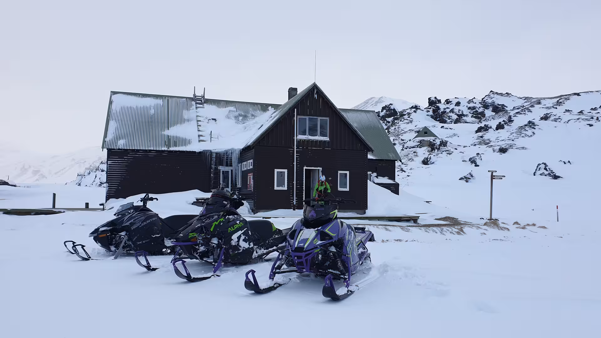 Two Arctic Cat snowmobiles parked outside a dark wooden mountain hut in Landmannalaugar Highlands covered in snow