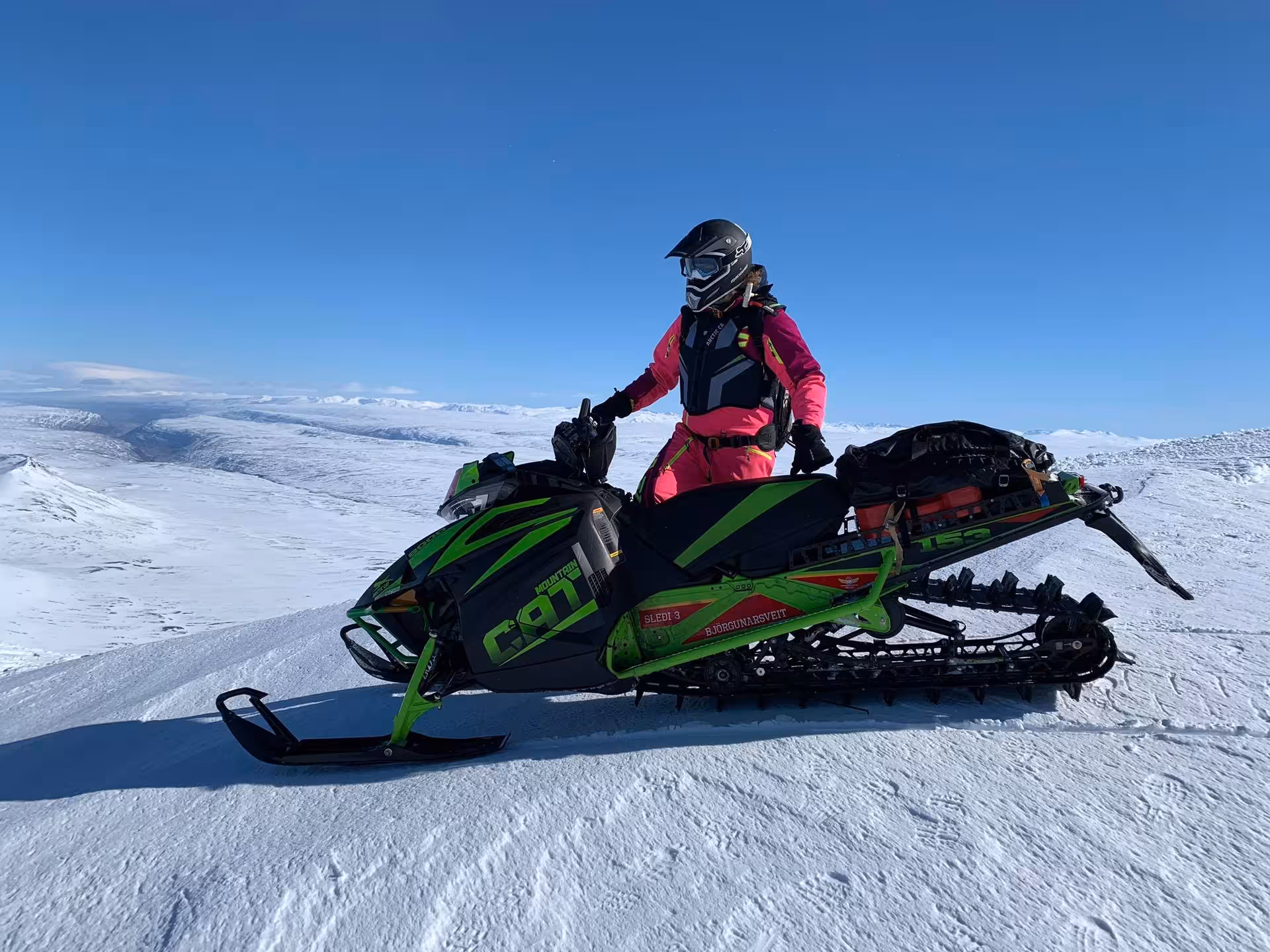 Rider in pink gear on green Arctic Cat snowmobile in snowy Landmannalaugar Highlands under blue sky
