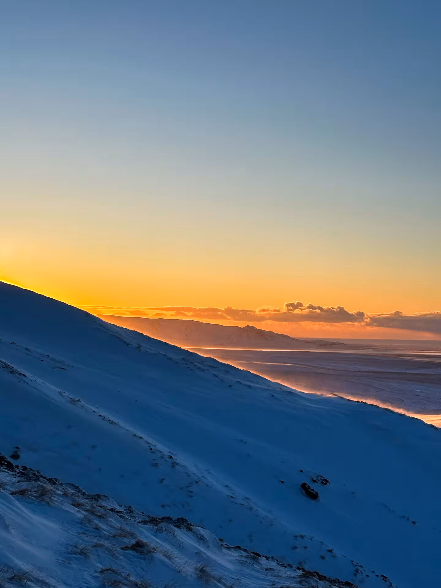 Snow-covered mountain slope at golden sunset with coastal views below clear blue sky