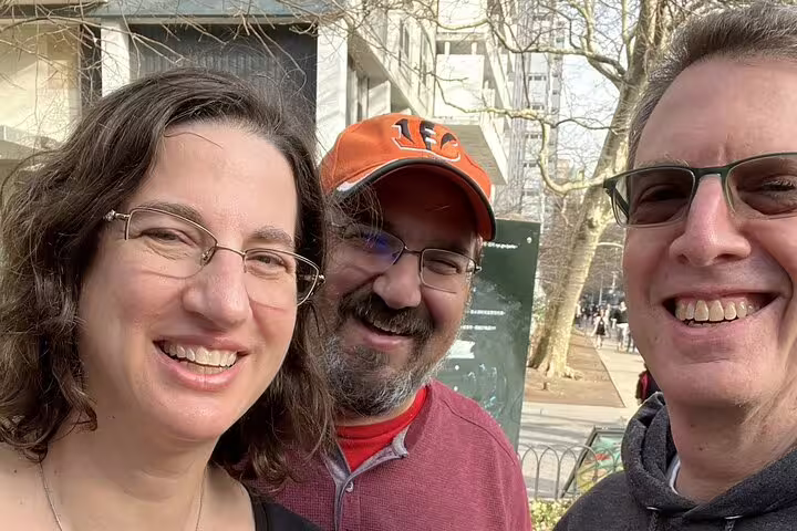 Three smiling adults posing for a selfie outdoors in an urban setting with bare trees and buildings.