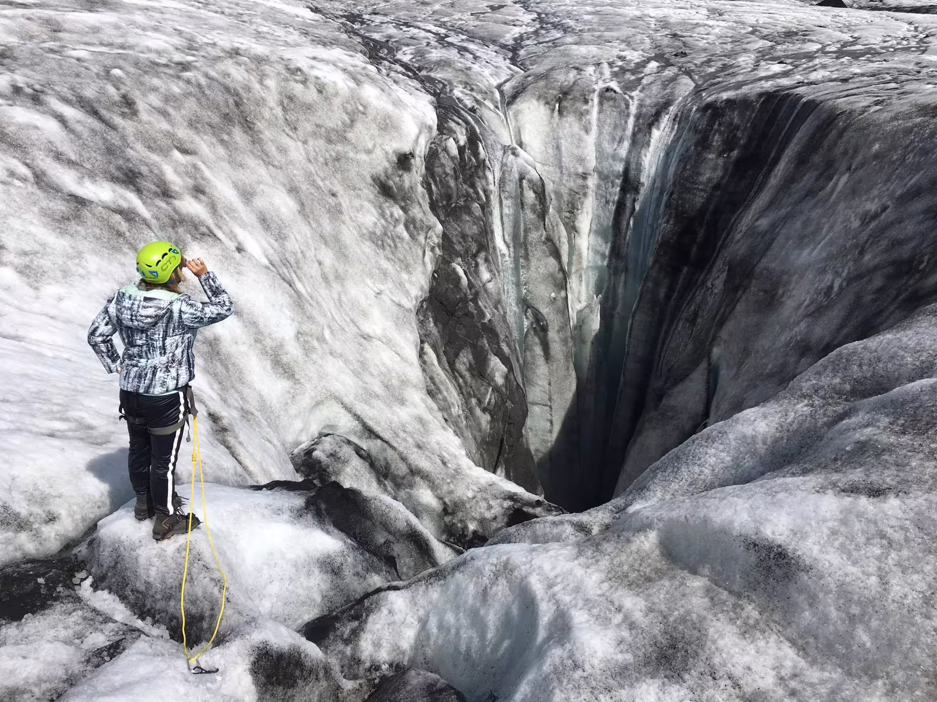 Hiker in green helmet peers into deep glacial moulin crevasse on Solheimajokull glacier, Iceland