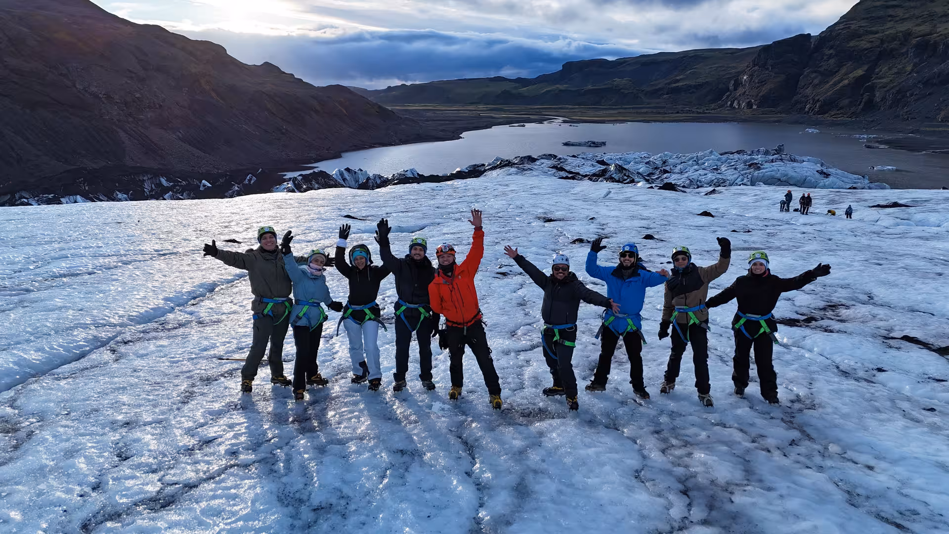 Group of tourists with harnesses and helmets posing on Solheimajokull glacier with glacial lagoon behind