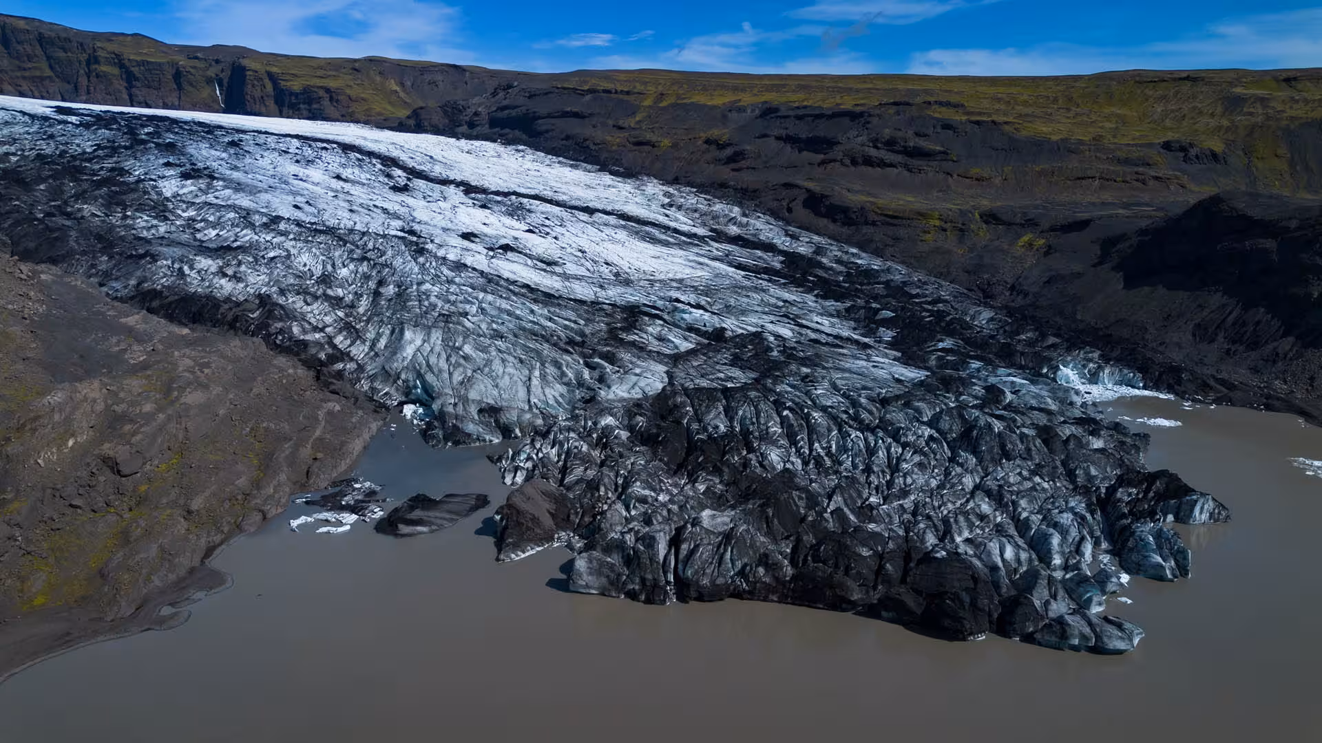 Aerial view of Solheimajokull glacier tongue meeting glacial lagoon with dark volcanic rock in Iceland