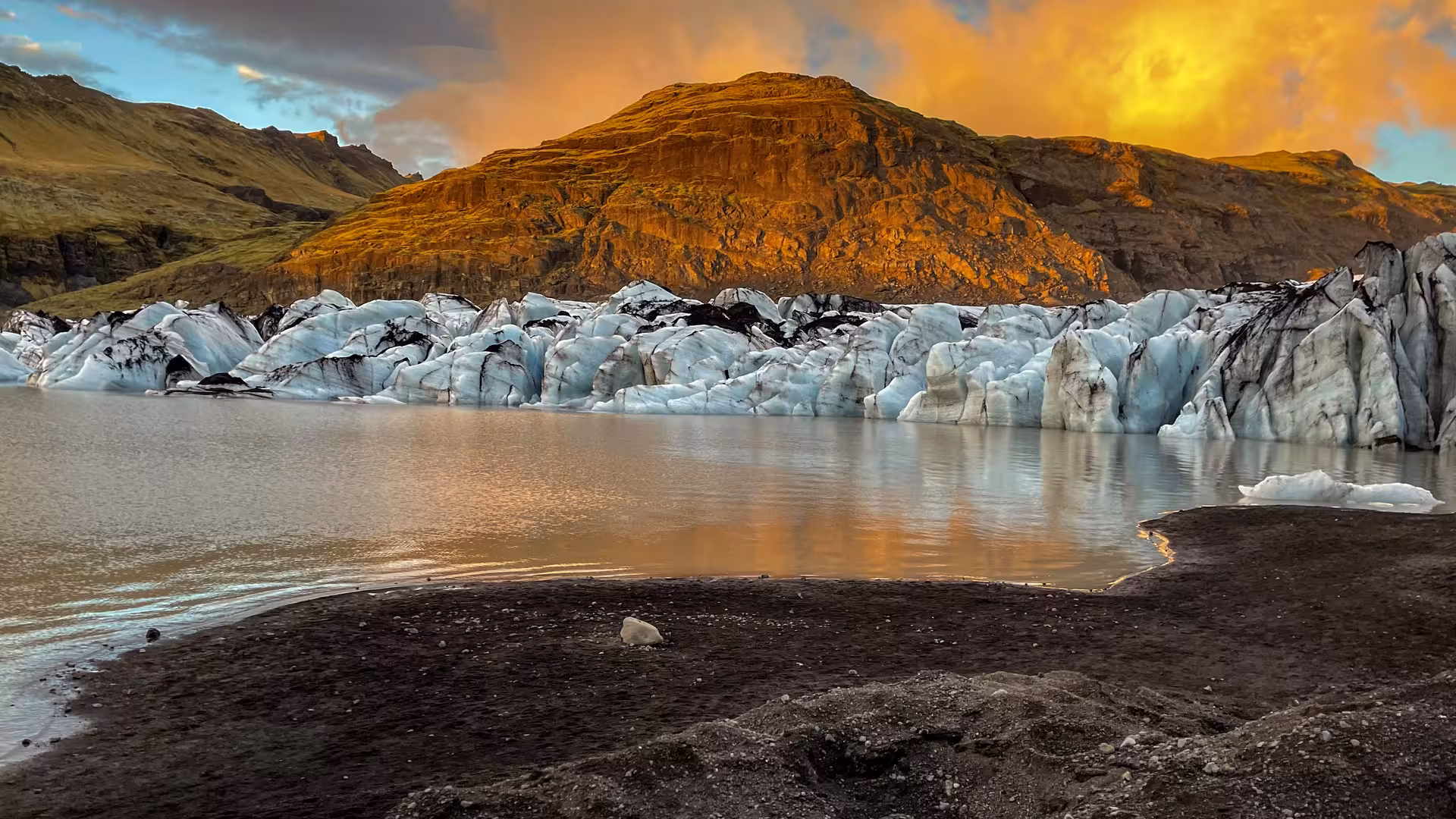 Solheimajokull glacier ice formations reflecting in glacial lagoon at sunset with volcanic mountain backdrop, Iceland