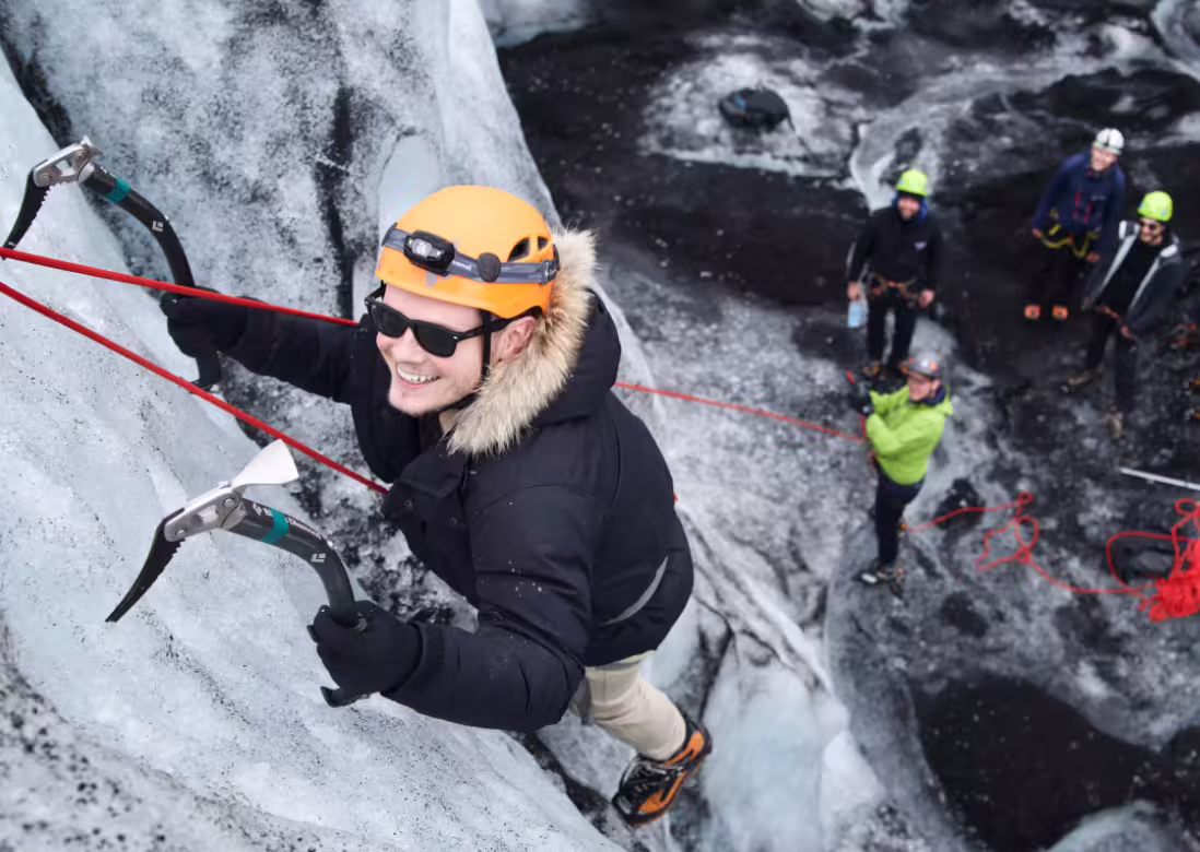 Smiling climber with ice axes and orange helmet scaling glacier wall at Solheimajokull with group below
