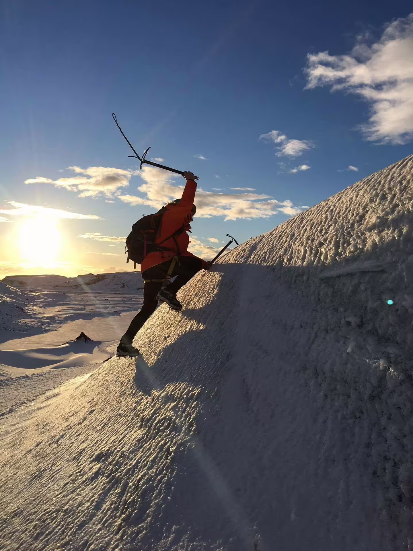 Climber in red jacket using ice axes to scale snowy glacier slope at sunset on Solheimajokull