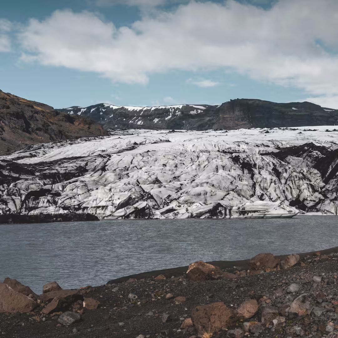 Panoramic view of Solheimajokull glacier meeting a glacial lagoon with rocky volcanic shoreline on a South Iceland guided tour.
