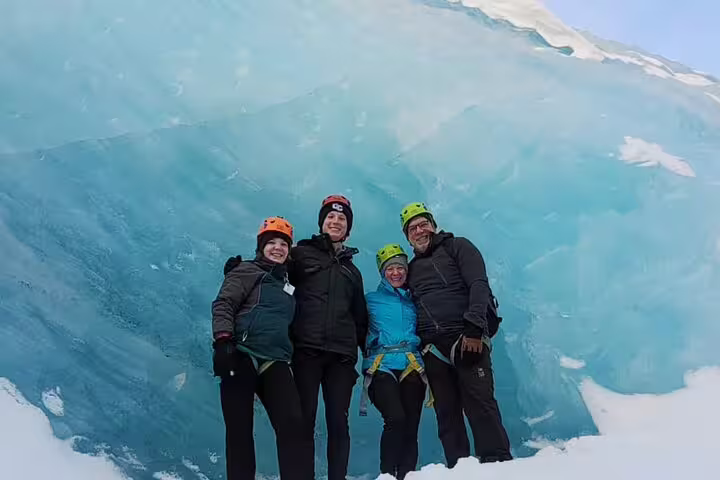 Four smiling hikers in helmets posing inside a blue ice cave on Sólheimajökull glacier, Iceland
