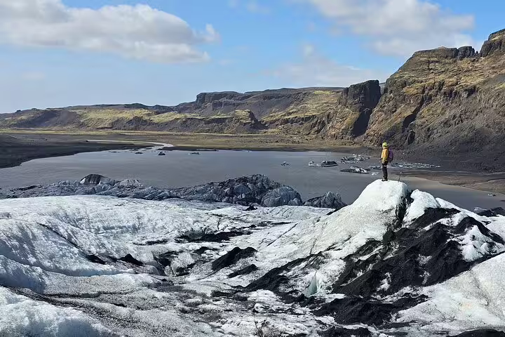 Hiker in yellow jacket standing on Sólheimajökull glacier with glacial lagoon and volcanic cliffs in background