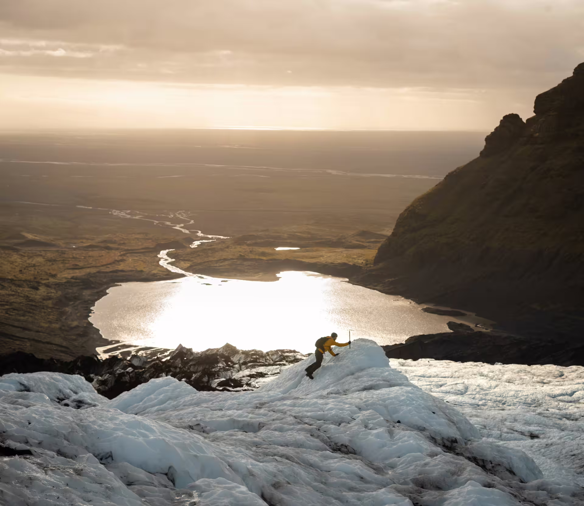 Solo hiker with ice axe climbs Iceland glacier at golden hour with breathtaking panoramic valley and lake views below