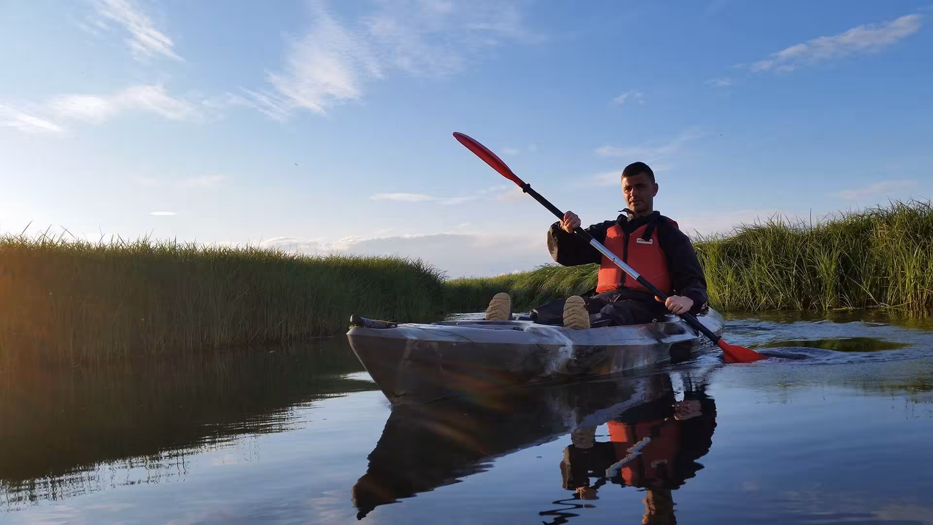 Man kayaking solo through narrow marsh waterway with red paddle and life vest on calm water