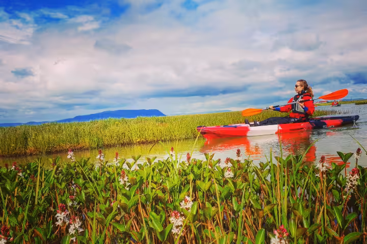 Woman kayaking solo through lush wetland marshes with red kayak and mountain backdrop under cloudy sky