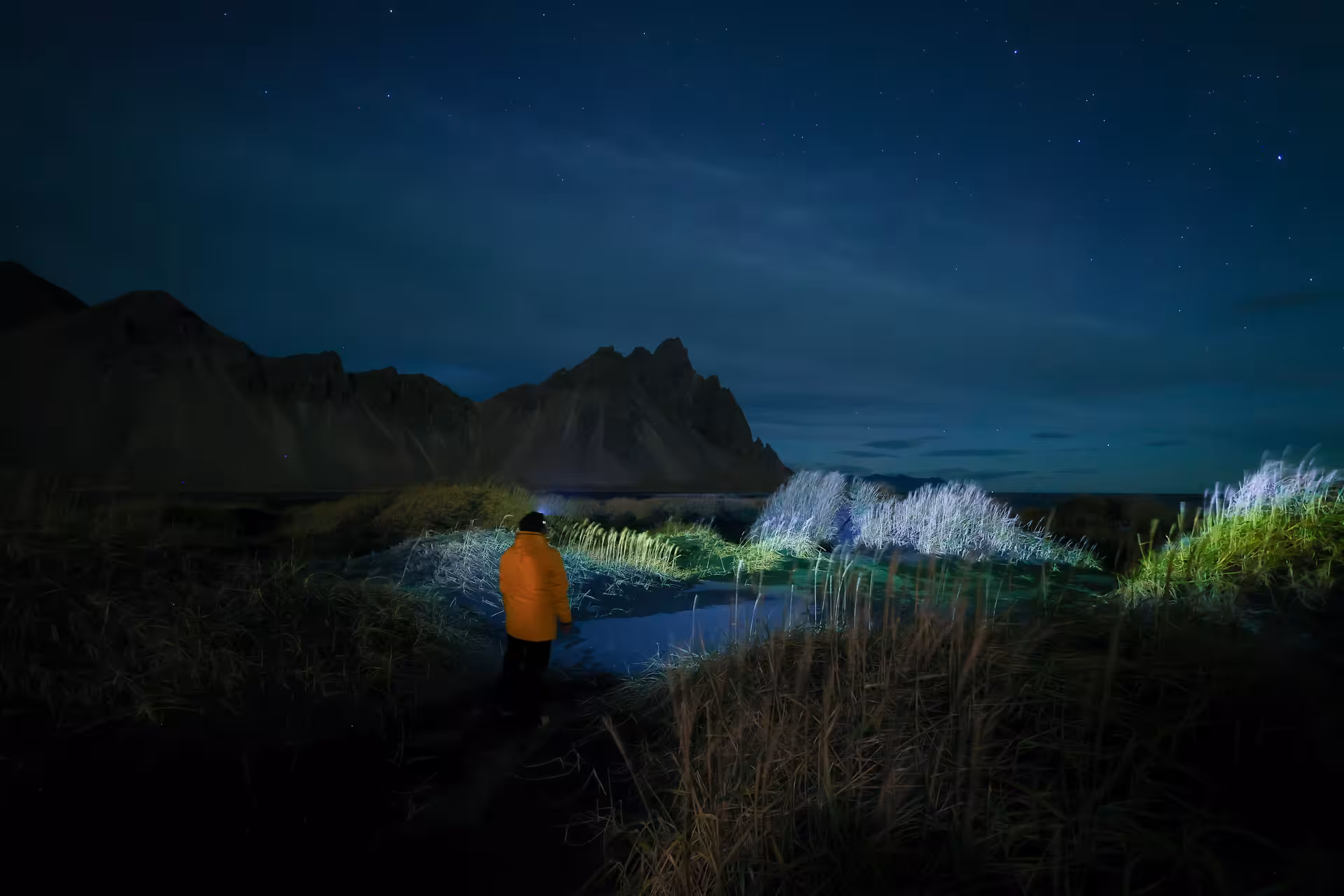 Solo traveler in orange jacket explores Iceland's dark wilderness landscape under a stunning starry night sky.
