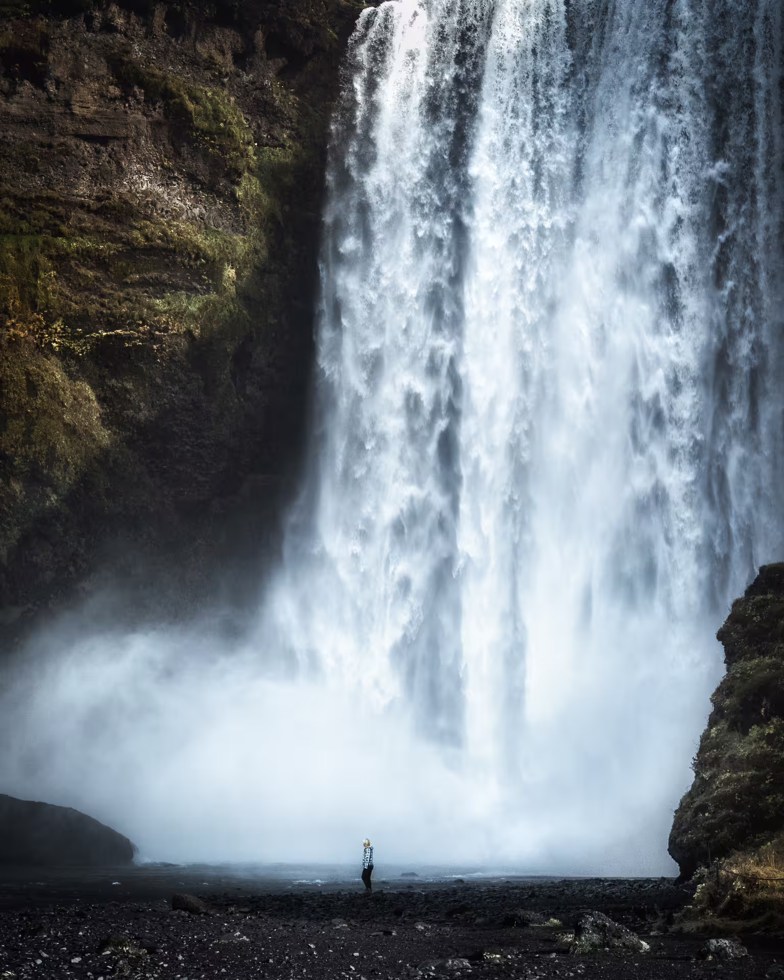 Solo traveler dwarfed by powerful Skogafoss waterfall mist on Iceland South Coast glacier tour