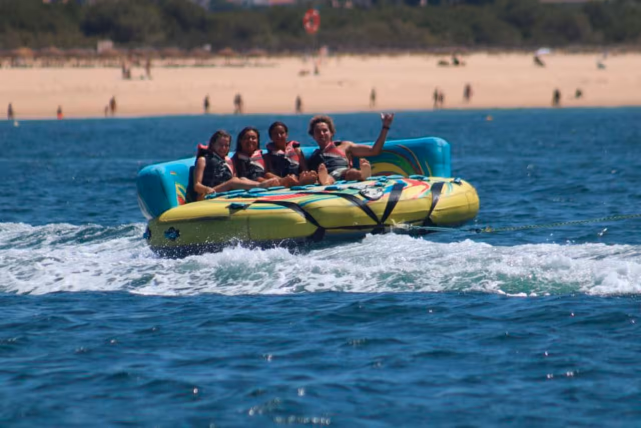 Friends relaxing on Sonar towable tube adventure, cruising behind a speedboat off a sunny sandy beach coastline