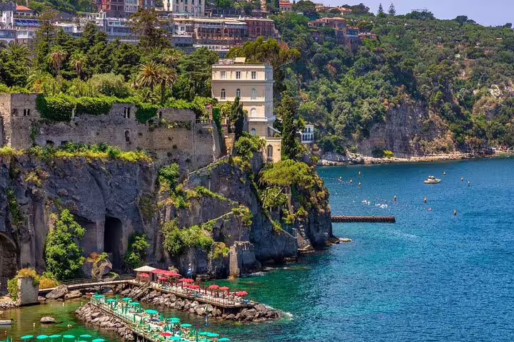 Cliffside view of Sorrento coastline with turquoise water, lush greenery, and colorful beach umbrellas below.