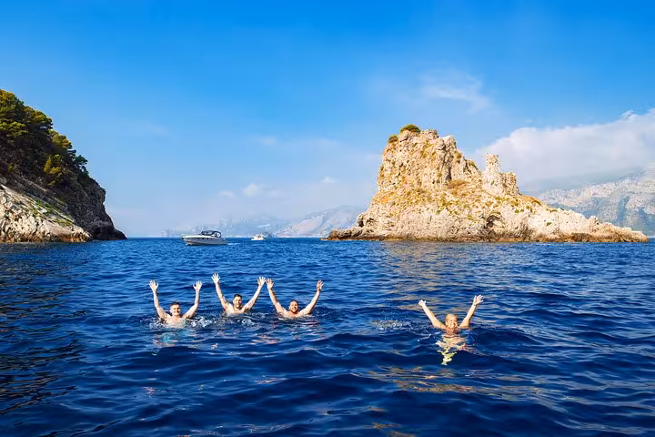 Four people swimming joyfully in the clear blue waters near rocky outcrops on a Sorrento Coast boat tour.