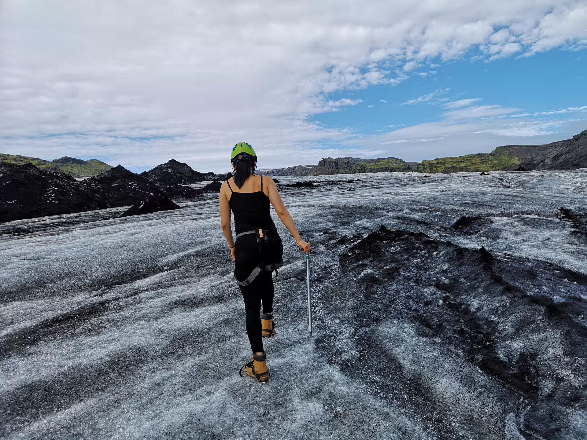Woman with ice axe and helmet hiking on a dark glacier with green mountains in Iceland