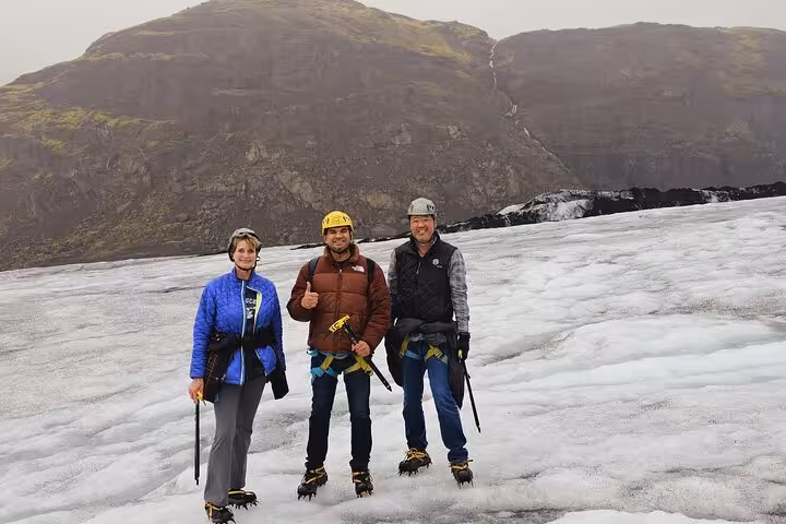 Three hikers with crampons and ice axes posing on a glacier with rocky mountain backdrop in Iceland