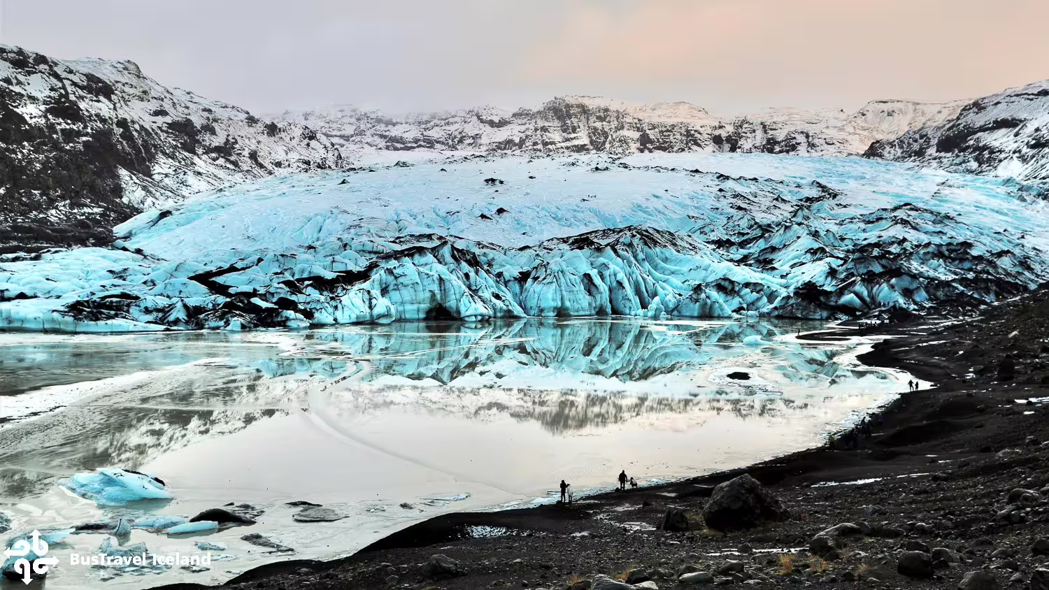 Brilliant blue Sólheimajökull glacier reflecting in glacial lagoon with tiny visitors on black sand shore, Iceland