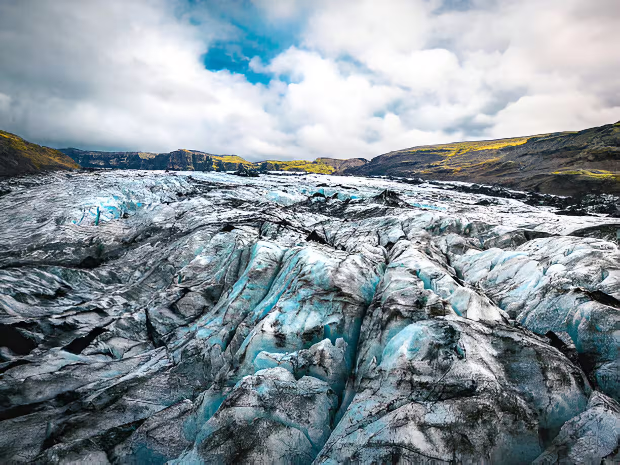 Cracked blue glacial ice surface with dark volcanic ash streaks and green mountains in background, Iceland