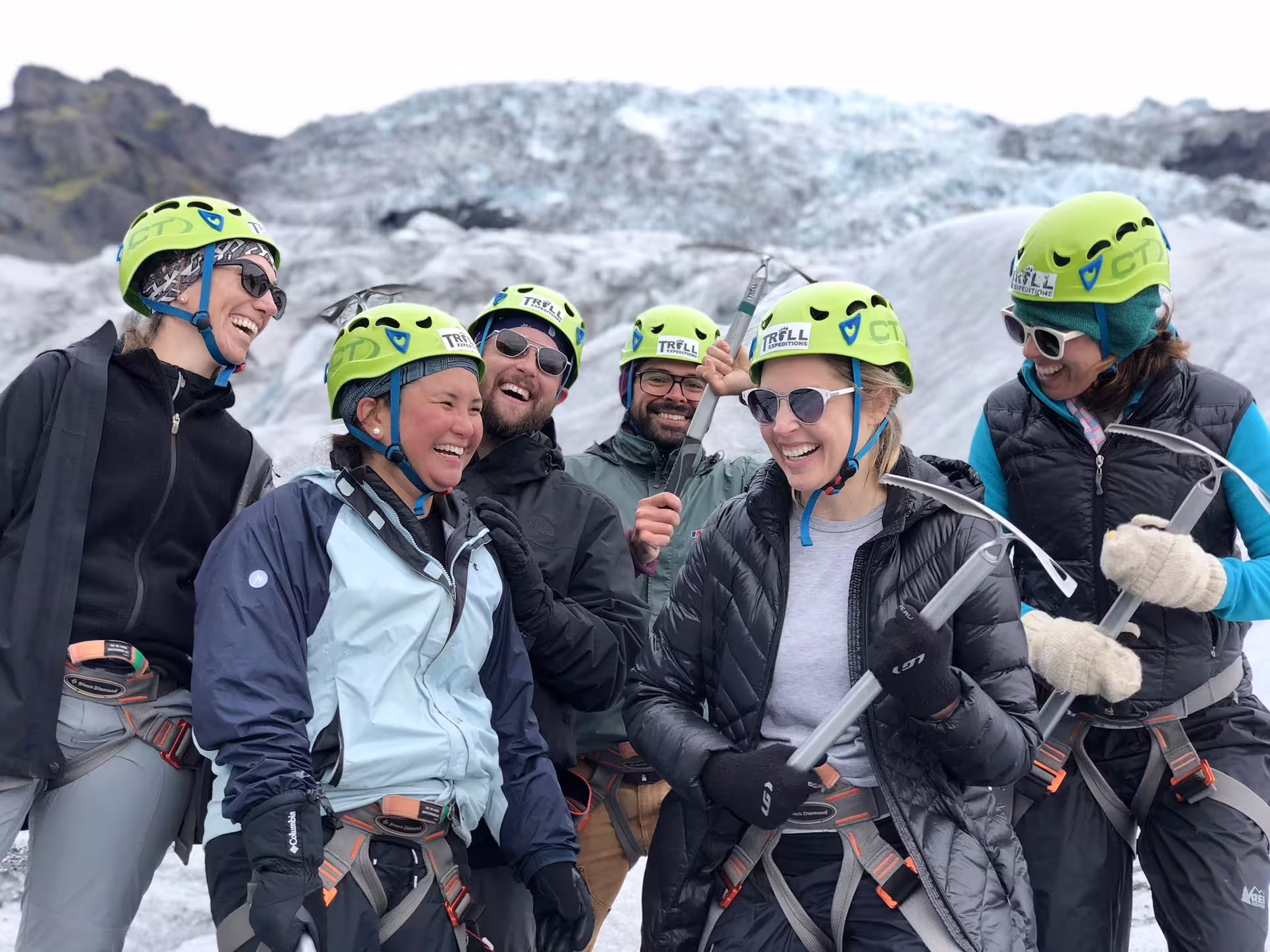 Group of smiling hikers wearing green Troll Expeditions helmets and harnesses on an Iceland glacier with ice axes