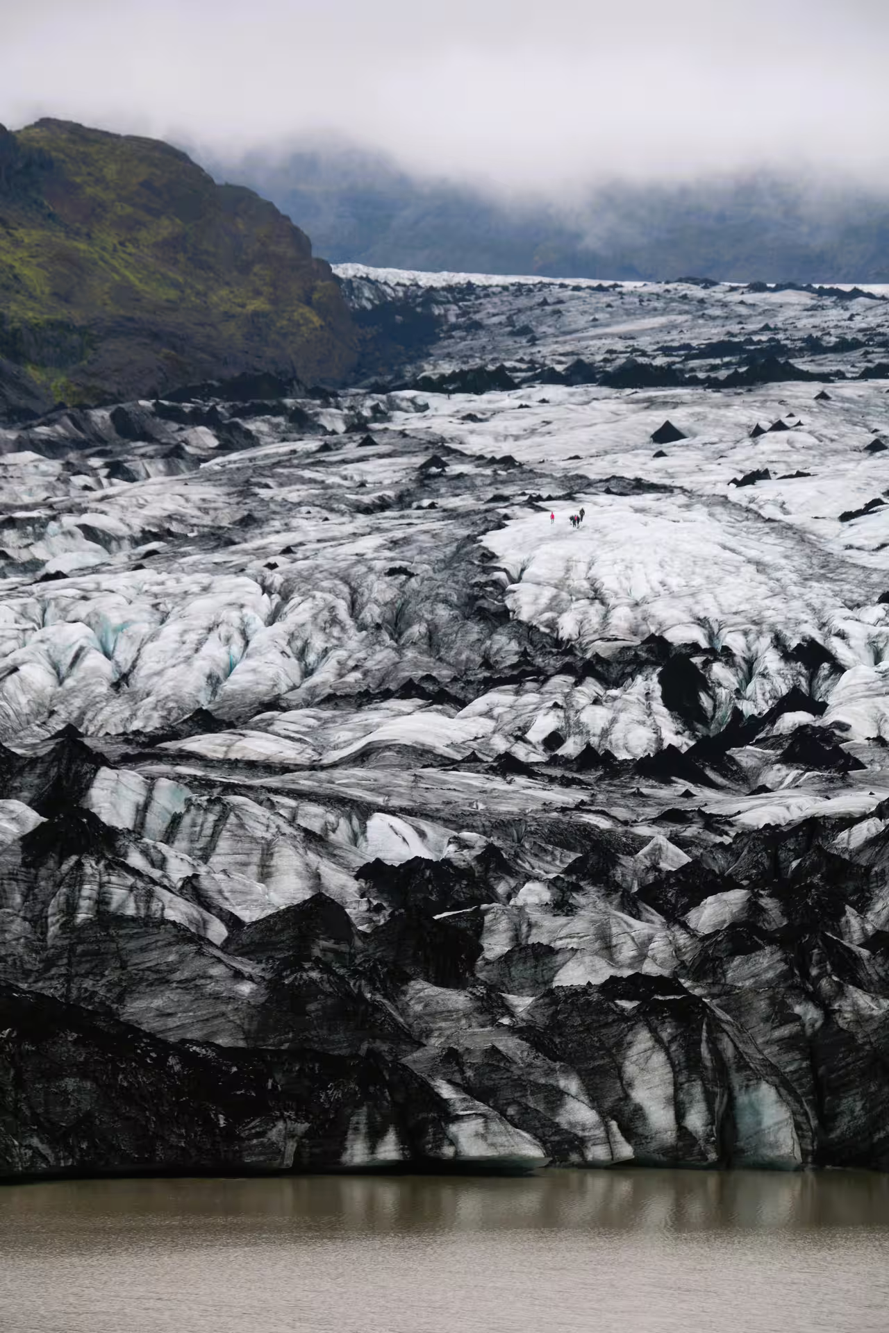 Hikers on a dark ash-streaked glacier with crevasses and glacial lake in Iceland's South Coast
