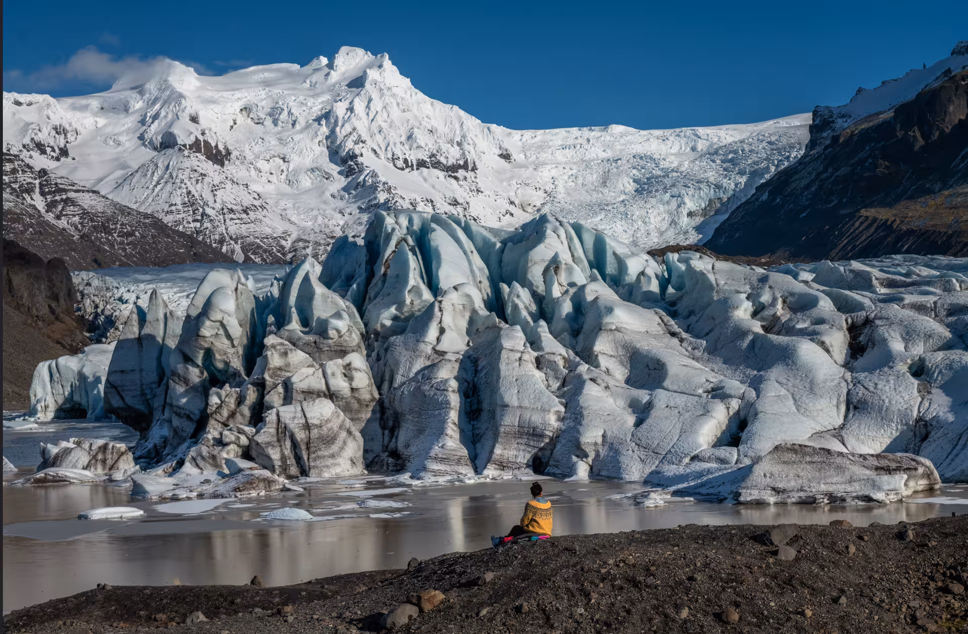 Person sitting beside a glacial lagoon with towering ice formations and snow-capped mountains in Iceland