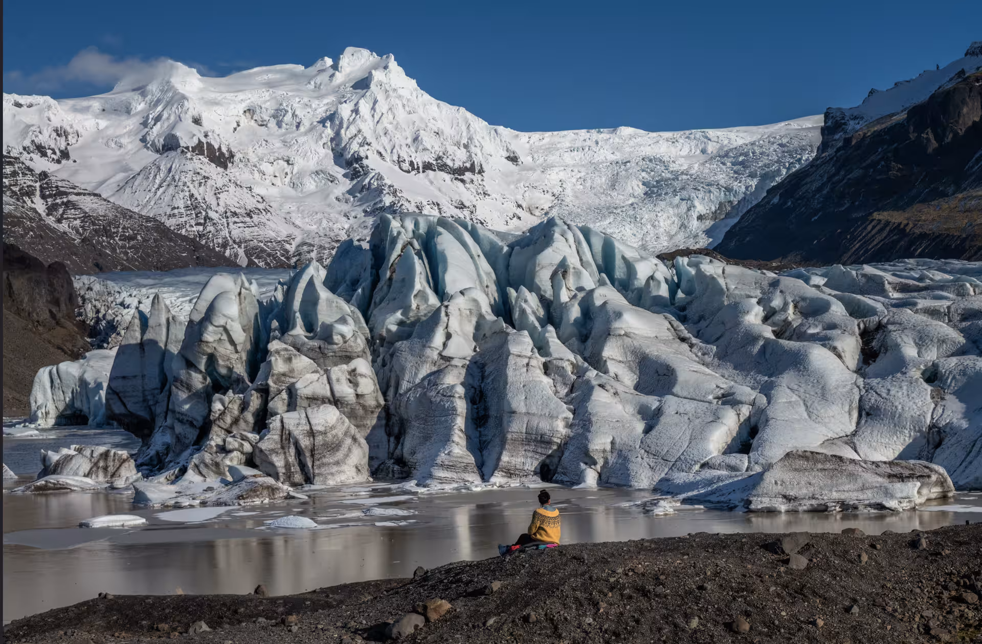 Person sitting beside a massive glacier with snow-capped mountains on Iceland's south coast