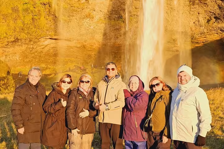 Group of seven tourists posing in front of Seljalandsfoss waterfall with rainbow on Iceland South Coast tour