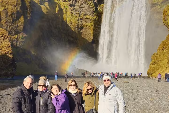 Group of six tourists smiling in front of Skógafoss waterfall with rainbow on Iceland South Coast tour