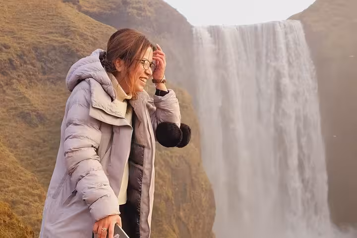 Smiling woman in winter coat standing before Skógafoss waterfall on Iceland South Coast tour