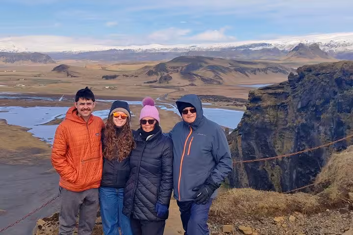 Four tourists posing on a cliff edge with Iceland's vast south coast valley and snow-capped mountains behind them